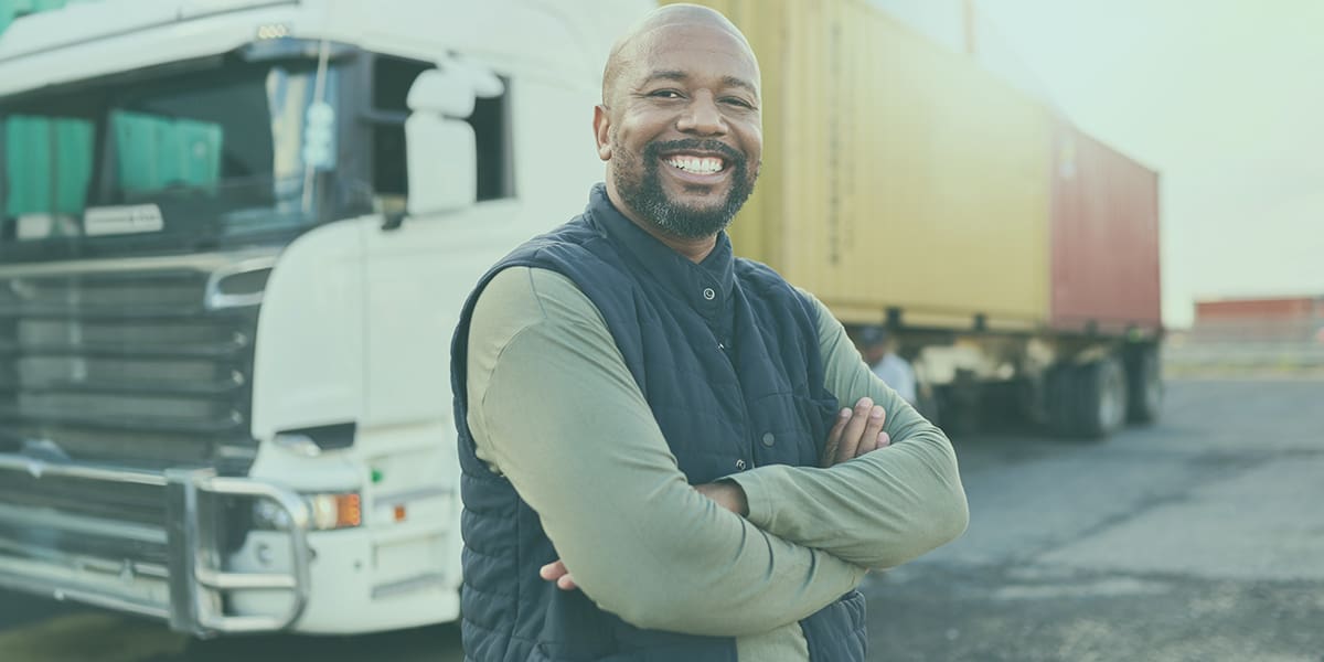 A truck driver standing in from of his transport truck with his arms crossed, smiling.