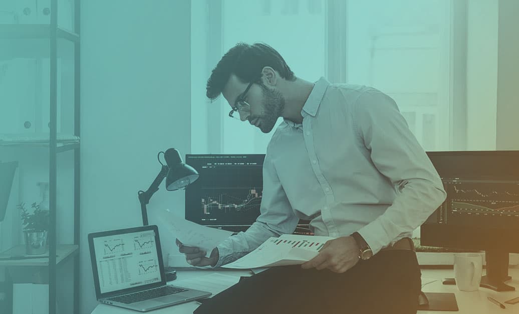 Business executive surrounded by computer monitors reviews data graphs printed on paper